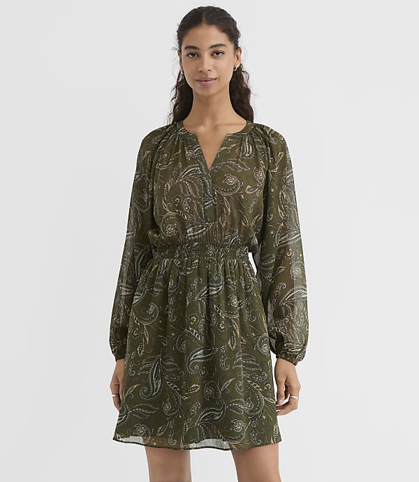 A woman in a green paisley print dress stands against a plain white background.