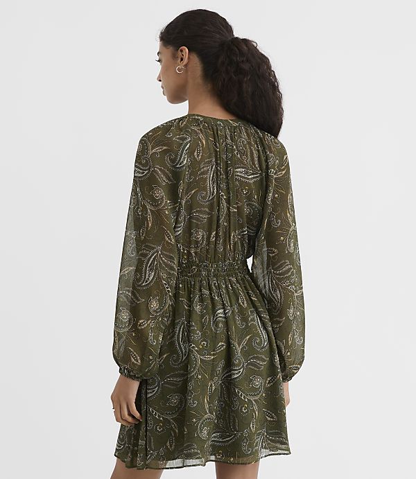 A woman in a green paisley dress with long sleeves stands against a plain, light background.