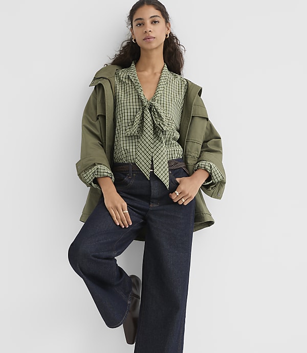 A woman in a green checkered blouse and dark jeans leans against a white wall, wearing an olive green jacket.