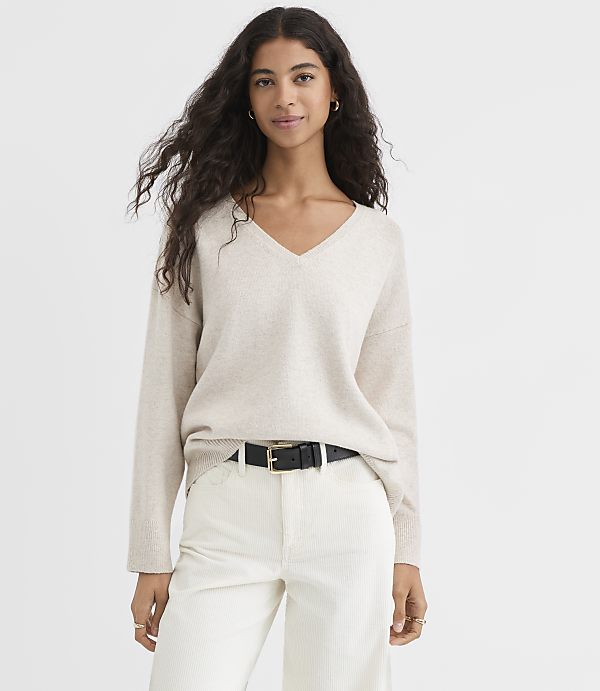 A woman in a beige v-neck sweater and white pants stands against a plain white background.