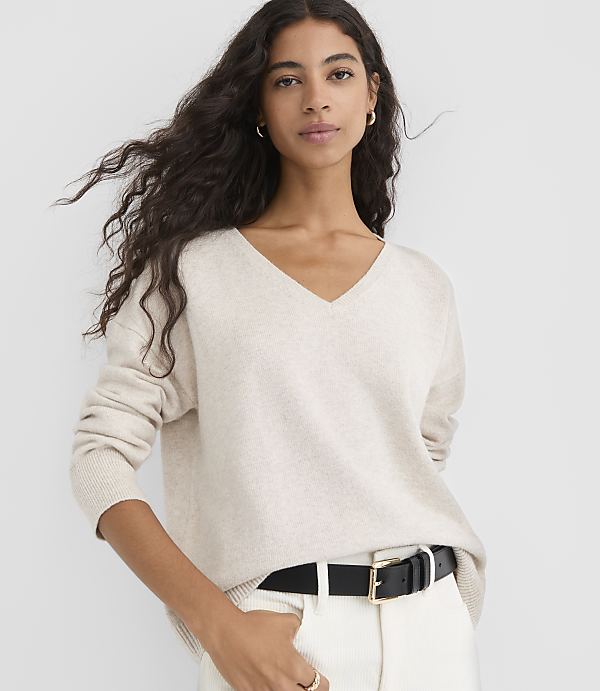 A woman in a cream v-neck sweater and white pants with a black belt stands against a plain white background.
