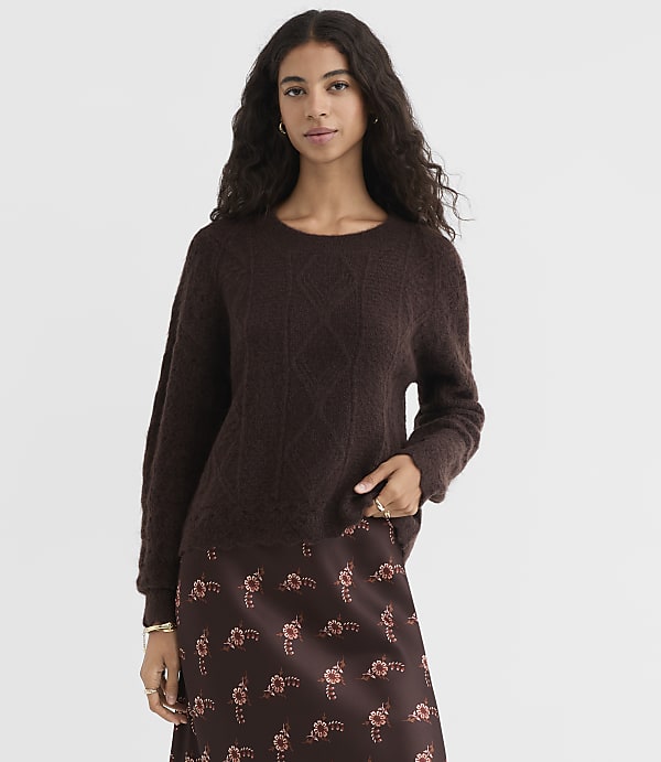 A woman in a dark brown cable-knit sweater and a floral print skirt stands against a plain, light brown background.