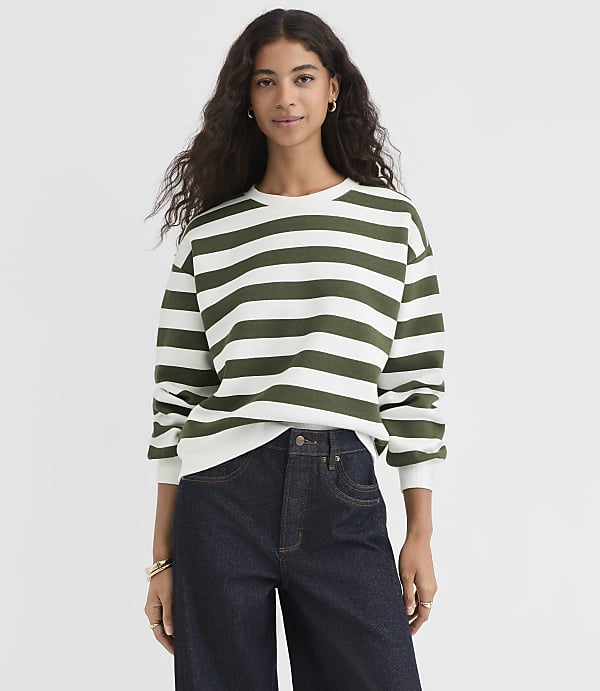 A woman in a green and white striped sweatshirt and dark jeans stands against a plain white background.