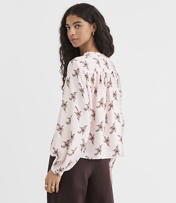A woman in a light pink, floral-patterned blouse and dark brown pants stands against a plain, light brown background.
