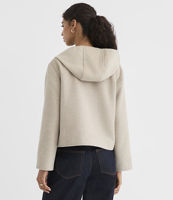 A woman in a beige hooded jacket and dark jeans stands against a plain white background.