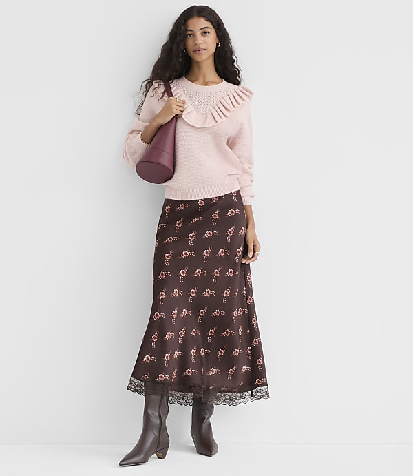 A woman in a pink ruffled sweater and a floral skirt stands against a plain white background.
