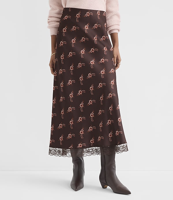 A woman in a floral print midi skirt and brown boots stands against a plain white background.