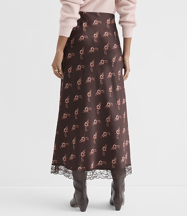 A person wearing a brown floral midi skirt with lace trim and a pink sweater stands against a plain white background.