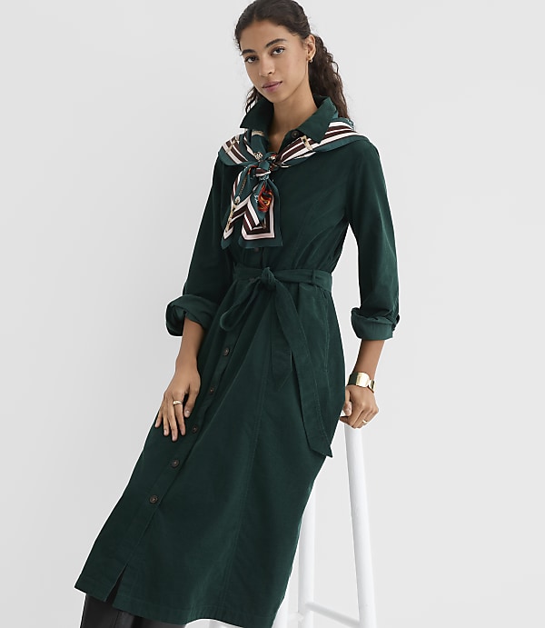 A woman in a green corduroy dress and patterned scarf sits on a white stool against a plain white background.