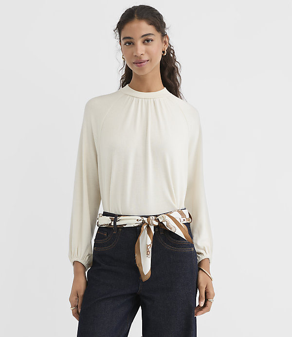 A woman in a cream-colored mock-neck top and dark jeans stands against a plain white background, with a patterned scarf tied around her waist.