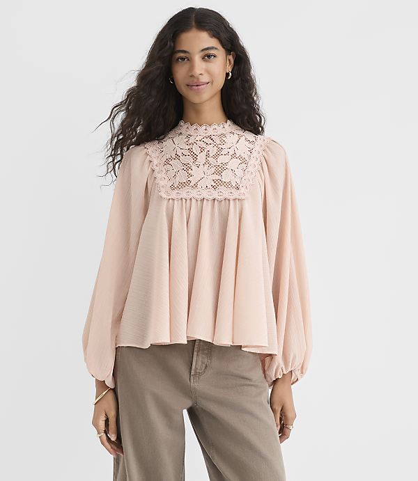 A woman in a light pink, lace-trimmed blouse and brown pants stands against a plain white background.
