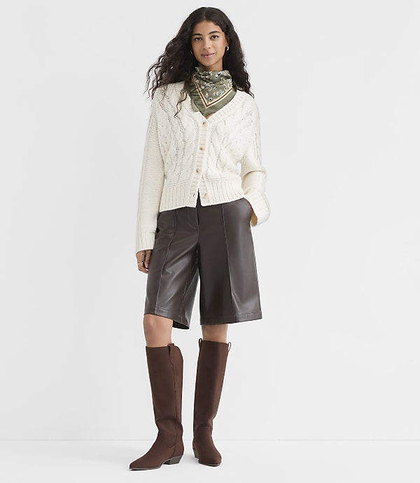 A woman in a cream cable-knit cardigan, brown leather culottes, and brown boots stands against a plain white background.