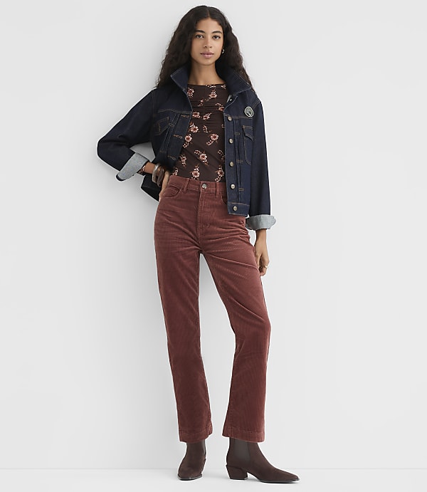 A woman in a denim jacket and rust-colored corduroy pants stands against a plain white background.