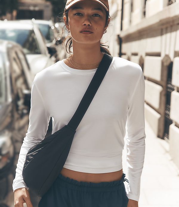 A young woman in a white long-sleeved shirt and a black crossbody bag stands on a city street with blurred cars and buildings in the background.