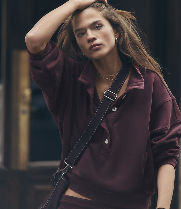 A woman in a maroon sweatshirt with a crossbody bag is captured with a blurred urban background.