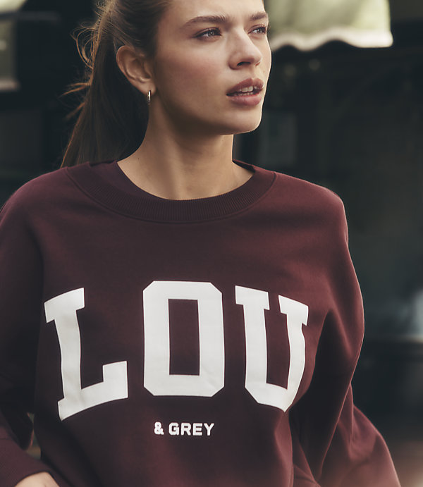 A young woman in a maroon sweatshirt with "LOU & GREY" printed on it is in the foreground, with a blurred outdoor background.