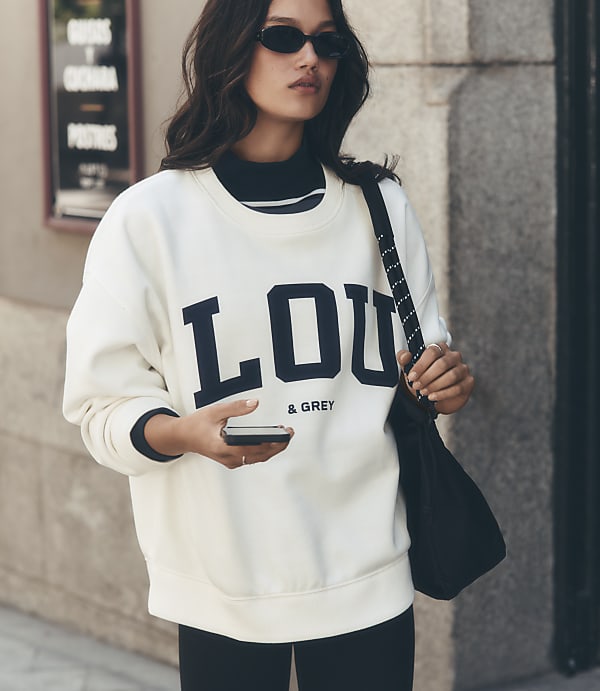A woman in a cream sweatshirt with "LOU & GREY" printed on it holds a phone in her hand while standing in front of a blurred urban background.