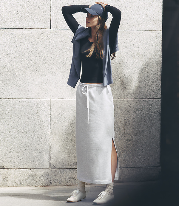 A woman in a black top, grey skirt, and blue cap stands against a textured stone wall.