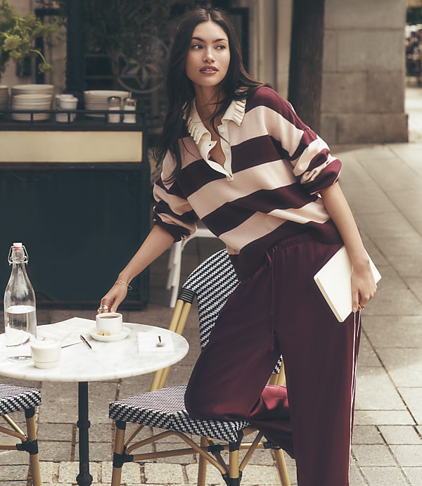 A woman in a striped shirt and maroon pants leans on a cafe table with a drink and book, while a blurred street scene with buildings and a sidewalk forms the background.