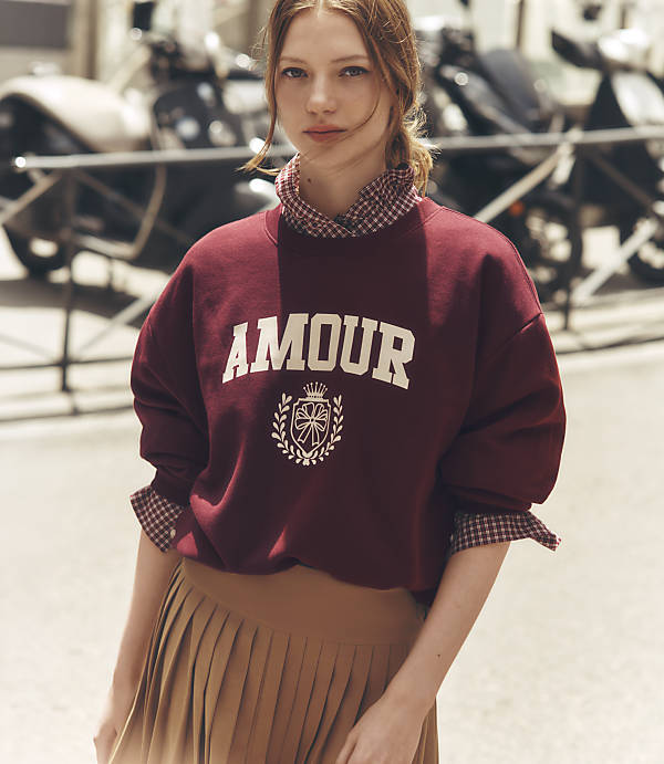 A young woman in a maroon sweatshirt with "AMOUR" written on it and a pleated skirt stands in front of a blurred background of parked motorcycles.