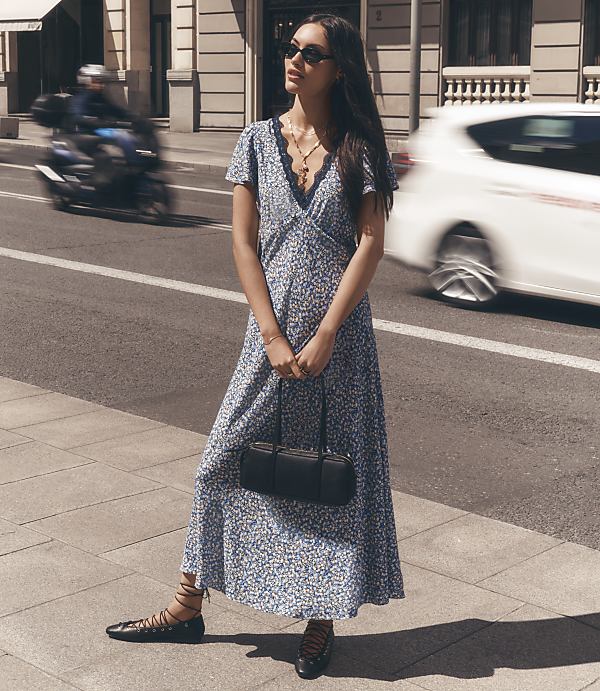 A woman in a floral dress stands on a sidewalk with a blurred motorcycle and car passing by in the background.