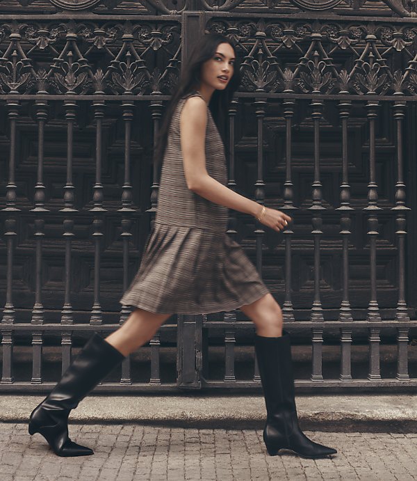 A woman in a plaid dress and black boots walks in front of an ornate metal gate.