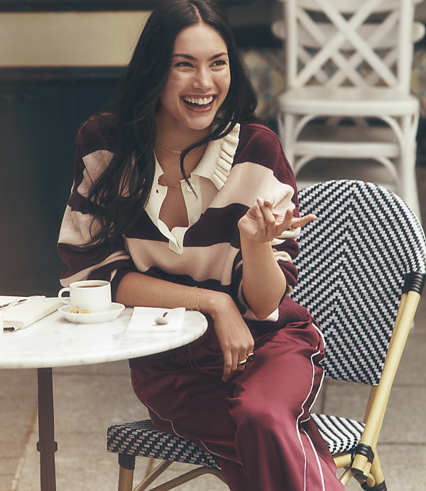 A laughing woman in a striped sweater and maroon pants sits at a small round table with a coffee cup, while a white chair is visible in the blurred background.