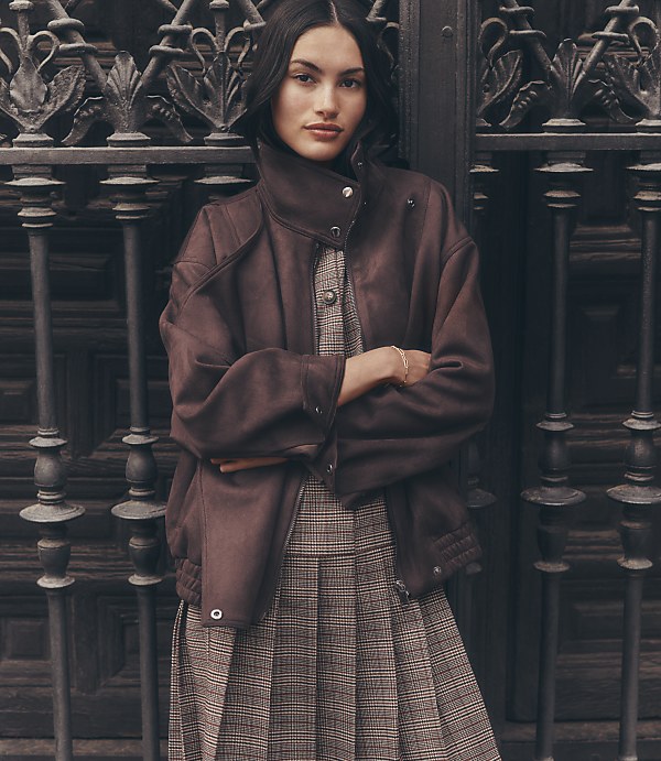 A woman in a brown jacket and plaid dress stands in front of an ornate metal gate.