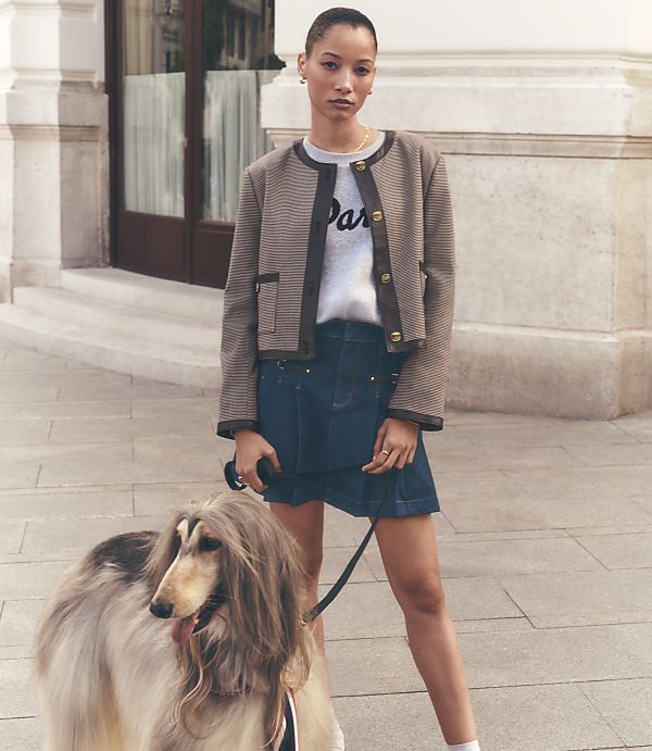 A woman in a stylish jacket and skirt stands with an Afghan hound on a sidewalk in front of a building.