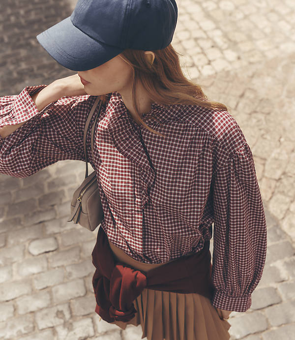A young woman in a gingham shirt and baseball cap walks on a cobblestone street, her pleated skirt visible.