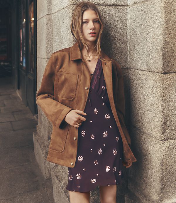 A young woman in a brown jacket and floral dress stands against a stone wall with a blurred street scene in the background.