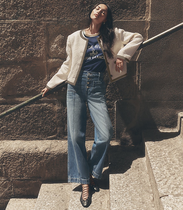 A woman in wide-leg jeans and a textured jacket poses on stone steps against a rough stone wall.