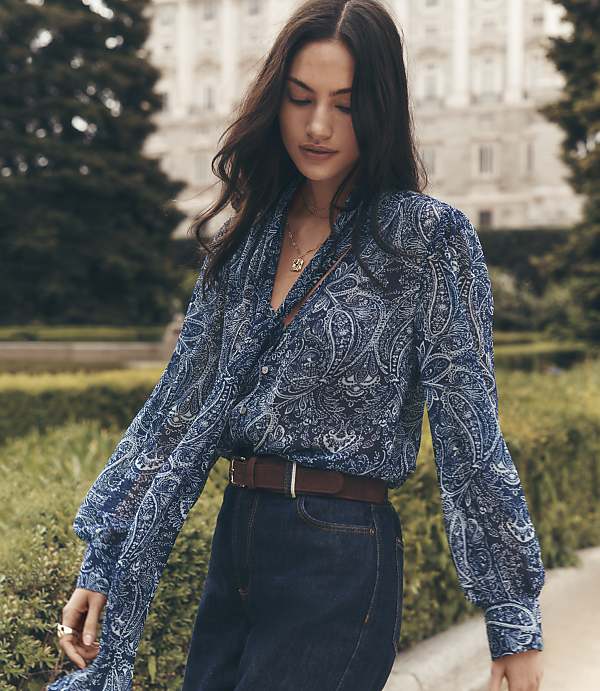 A woman in a blue paisley blouse and dark jeans stands in front of a grand building with trees.