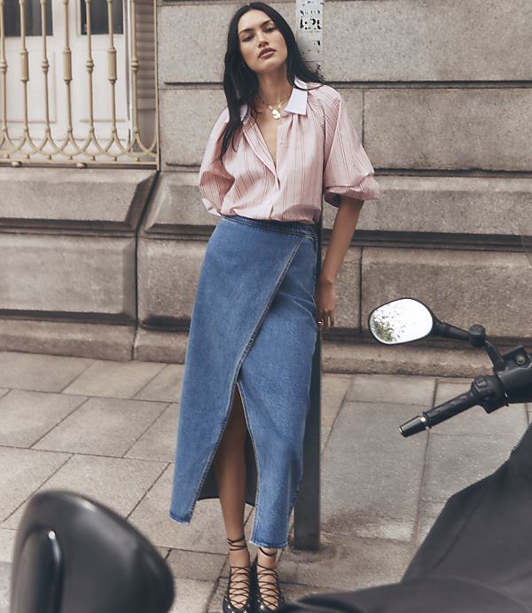 A woman in a pink striped shirt and denim skirt leans against a metal barrier with blue arrows, while a parked motorcycle and a building are visible in the blurred background.