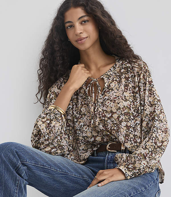 A woman with long, dark, curly hair wears a floral print blouse and blue jeans, seated against a plain, light-colored background.