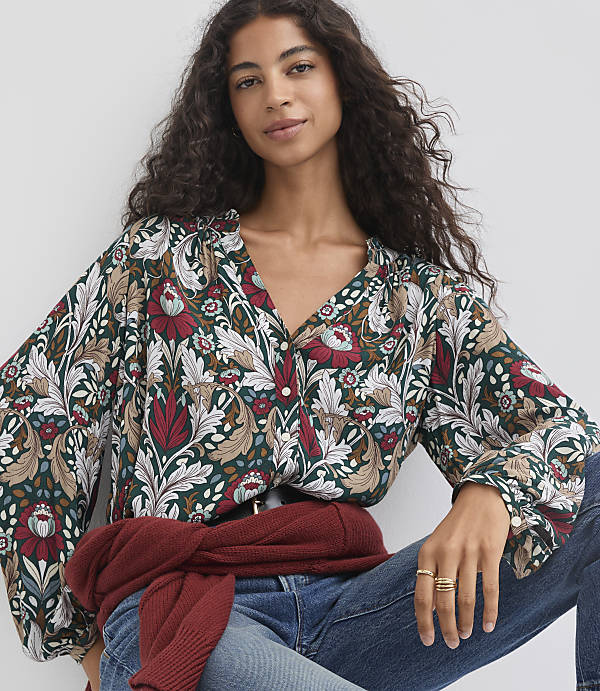 A woman with dark curly hair wears a dark green floral blouse and blue jeans, with a maroon sweater tied around her waist, against a plain white background.