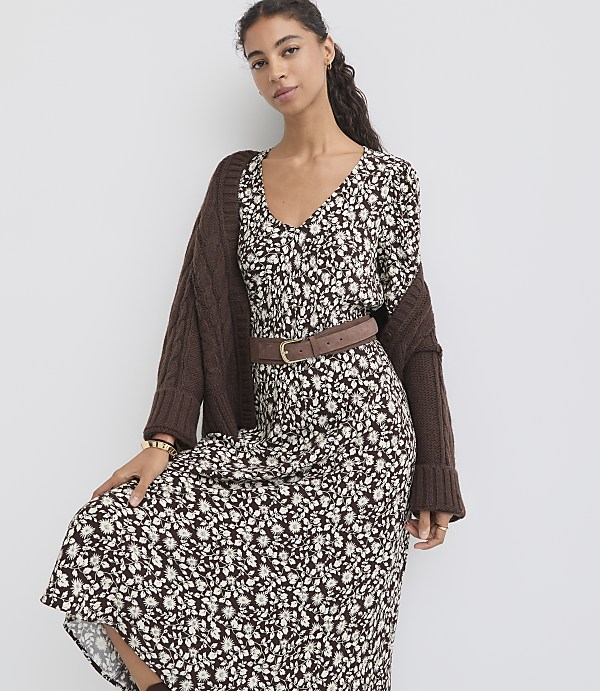 A woman in a floral dress and brown cardigan stands against a plain white background.