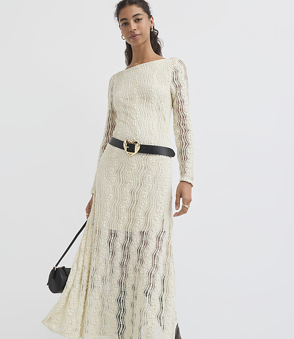 A woman in a cream-colored lace dress with a black belt and purse stands against a plain white background.