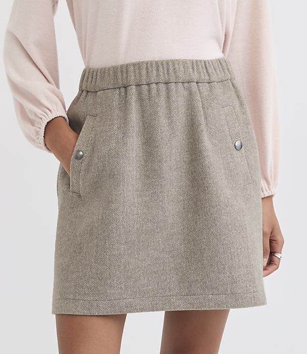 A person in a light pink top models a tan tweed mini skirt with pockets in the foreground against a plain white background.