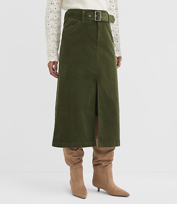 A person stands wearing a green corduroy skirt with a slit and a white patterned sweater, with tan boots peeking out from beneath the skirt, against a plain white background.