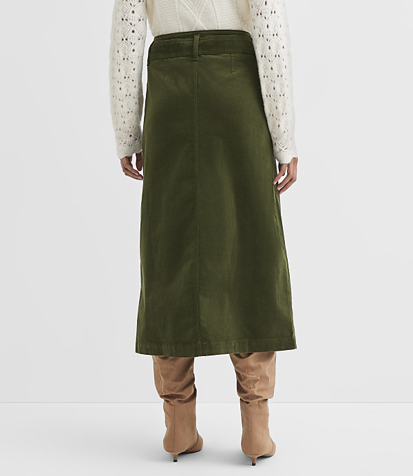 A person wearing a cream-colored sweater and a green corduroy skirt stands in front of a plain white background.