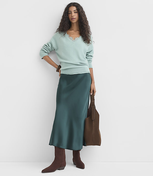 A woman in a light blue sweater and a teal satin skirt stands against a plain white background, holding a brown tote bag.