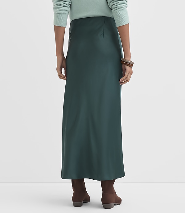 A person wearing a teal sweater and a dark green satin skirt stands against a plain white background, with brown boots visible at their feet.