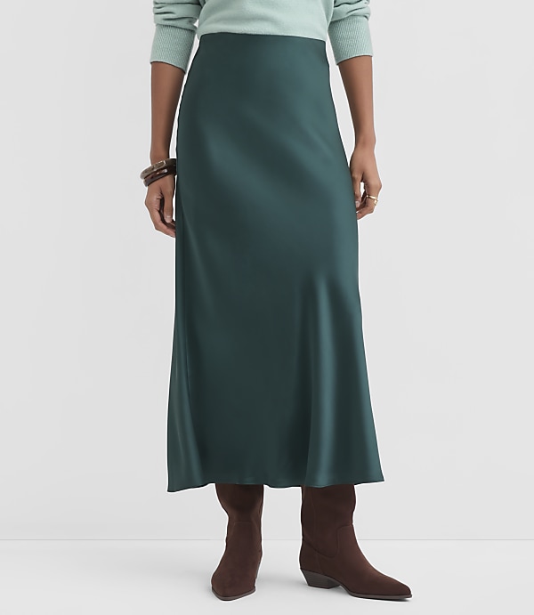 A woman in a teal sweater and a dark green satin skirt stands in front of a plain gray background, wearing brown boots and bracelets.