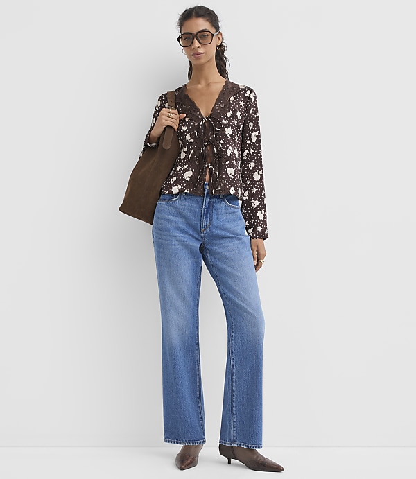 A woman in a floral blouse and jeans stands against a plain white background, carrying a brown tote bag.