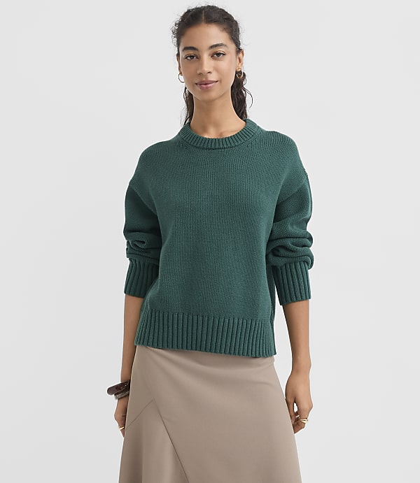 A woman in a teal sweater and tan skirt stands against a plain white background.