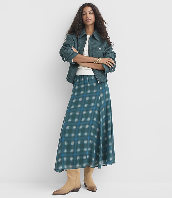 A woman in a teal tweed jacket and plaid skirt stands with her arms crossed against a plain white background.