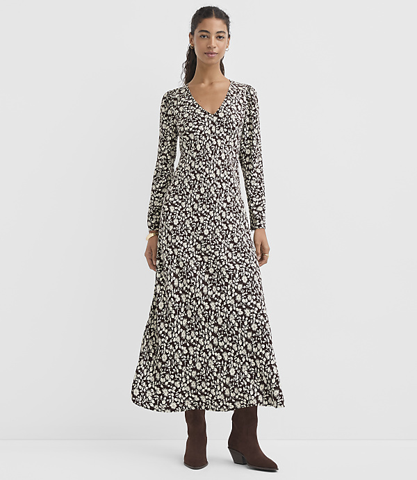 A woman in a long, patterned dress and brown boots stands against a plain, light gray background.