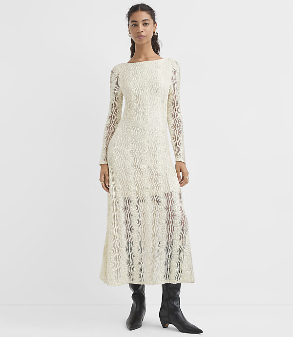A woman in a cream-colored, textured, long-sleeved dress and black boots stands against a plain white background.