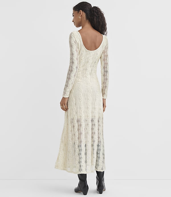 A woman in a cream-colored, long-sleeved, textured dress and black boots stands against a plain white background.